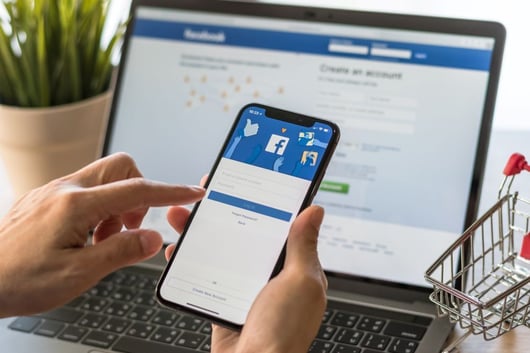 User logging into Facebook on a smartphone with a laptop in the background, showcasing b2b advertising platforms.